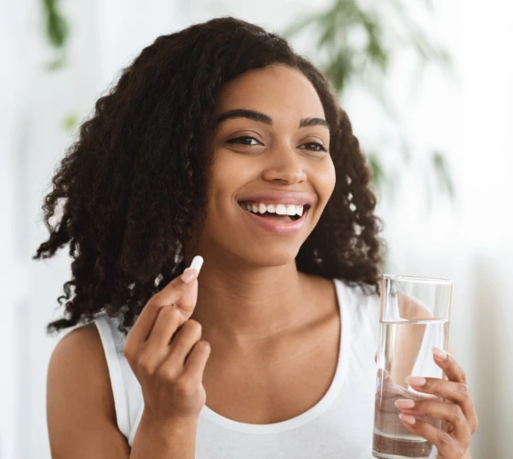 A smiling woman holds a glass of water in one hand and a pill in the other, preparing to take an oral dose. The image emphasizes health, wellness, and convenience, aligning with Aptar Pharma’s Activ-Polymer™ technology.