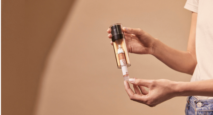 A person holds a dual-chamber airless named Neomix skincare product with a pump dispenser in one hand and removes a tubular insert from the bottom compartment, set against a neutral beige background.