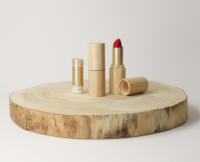 A red lipstick with a Non-Guided Lipstick Mechanism named Iconic and a lip balm in gold and bamboo tubes are displayed on a round, flat wooden slab against a plain white background.