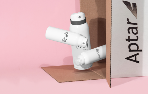 Two white aerosol cans labeled Glide by Aptar, and Apollo by Aptar are arranged on a pink background, partially inside a brown cardboard box with Aptar printed in bold black letters to highlight the design of  e-commerce capable aerosol products