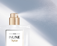 Close-up of a clear perfume bottle with a gold high definition spray fragrance pump releasing a fine mist. The label reads HDS INUNE by Aptar against a soft, light blue and white background.
