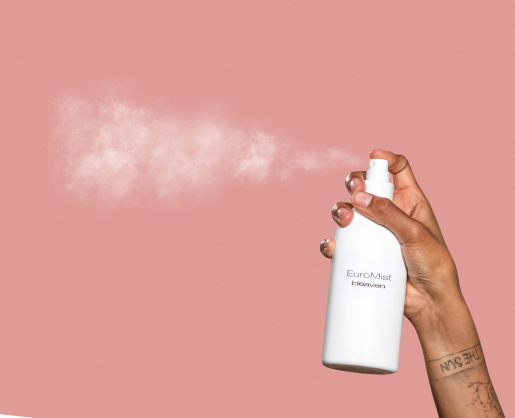 A hand with sprays a white bottle labeled EuroMist Heaven against a pink background, releasing a visible mist.