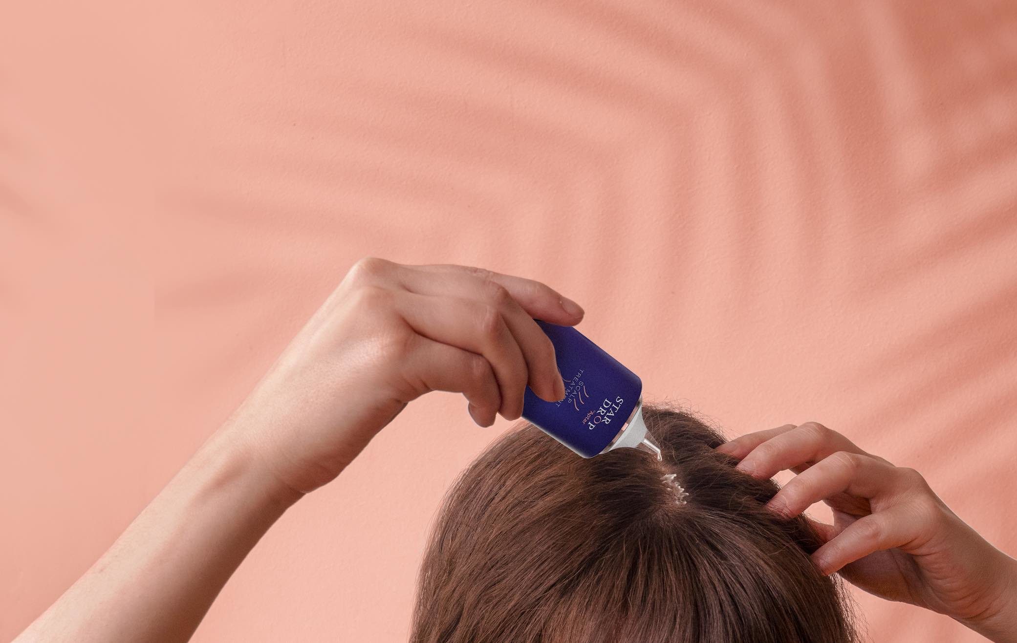 A person uses a small blue bottle with sleek haircare packaging to apply a liquid product directly onto their scalp, parting brown hair with one hand, set against a peach-colored background with a shadow pattern.