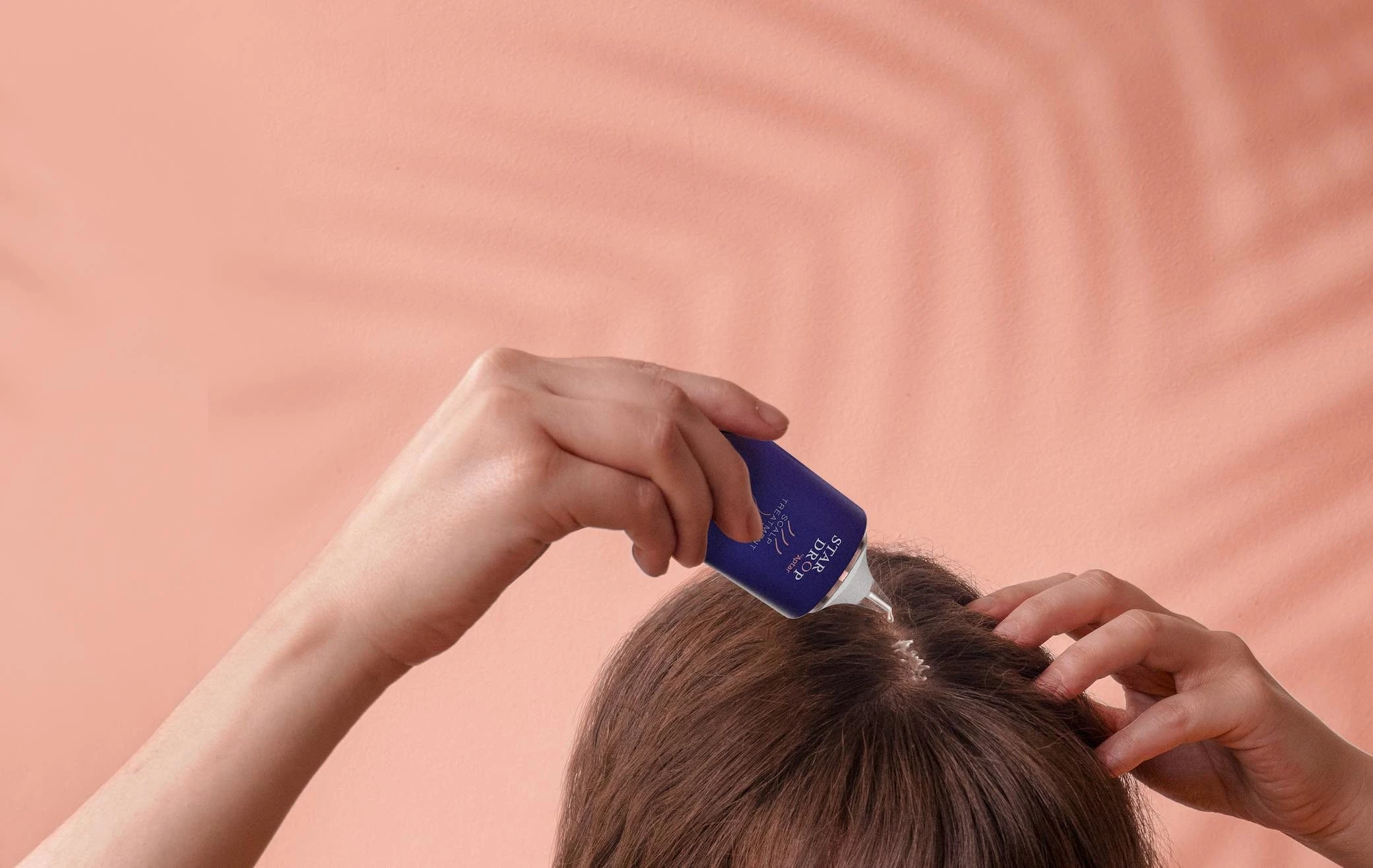 A person uses a small blue bottle with sleek haircare packaging to apply a liquid product directly onto their scalp, parting brown hair with one hand, set against a peach-colored background with a shadow pattern.