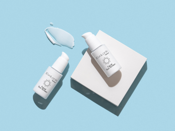 Two white pump bottles labeled Evolock facial sunscreen, featuring a twist-to-lock cosmetic pump  are displayed on a light blue background, one on a white square platform, the other beside a smear of light blue cream.
