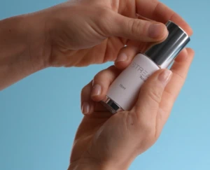 A person holds a small airless glass dispenser of cream or serum with a silver cap against a light blue background, preparing to open it. The bottle has the logo Astrea on it in silver to match the cap.