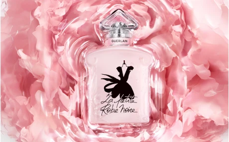 A high quality prestige fragrance pump labeled Sensea tops the Guerlain La Petite Robe Noire perfume bottle, which rests on soft, pink, petal-like swirls and showcases a black dress silhouette on the front.
