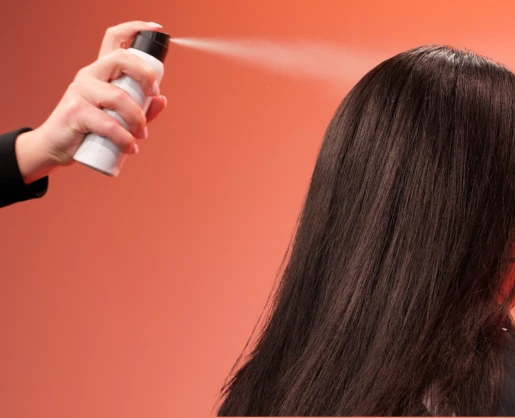 An image of a white aerosol spray can with a black actuator spraying dry shampoo on brown hair, depicting that a vertical slider aerosol valve is highly compatible with high powder formulations. The background is coral pink.