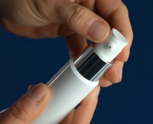 A close-up of hands holding and pressing the nozzle of a white and silver airless pump against a dark blue background.