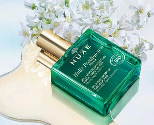 A green glass bottle of NUXE Huile Prodigieuse multi-purpose dry oil with a spray pump for dry oils named Oil Mist and gold cap, placed on a light reflective surface beside white flowers. The label displays product details and a certification badge.