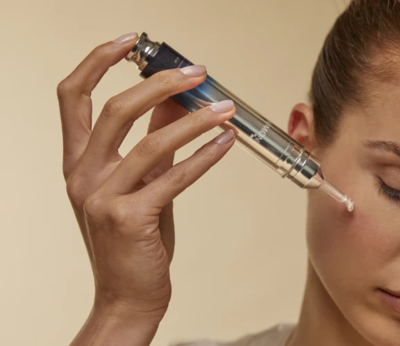 A person applies skincare serum under their eye using a metallic dropper syringe, showcasing innovative beauty packaging against a beige background. Only part of their face and hand are visible.