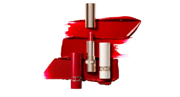 A red lipstick with a gold cap stands upright in front of two other lipsticks, one red and one white, showcasing bold red lipstick smears on a white background and highlighting its elegant refillable lipstick packaging.