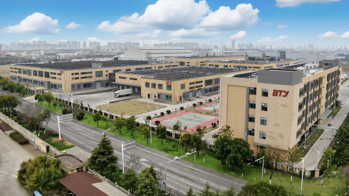 A large industrial complex with multiple tan buildings, green trees, a tennis court, solar panels on the roofs, and wide, empty roads, set under a blue sky with scattered clouds. In the image is BTY's manufacturing location in Huai'an.