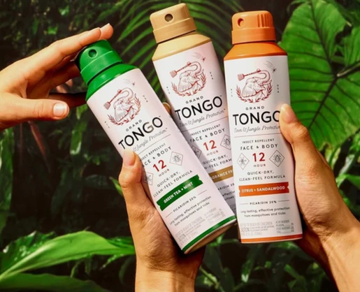 Three hands hold up three cans of Grand Tongo insect repellent spray with a premium twist to lock aerosol actuator named Markus, in a lush, green jungle setting. The green, yellow, and orange cans are labeled for face and body with different scents.