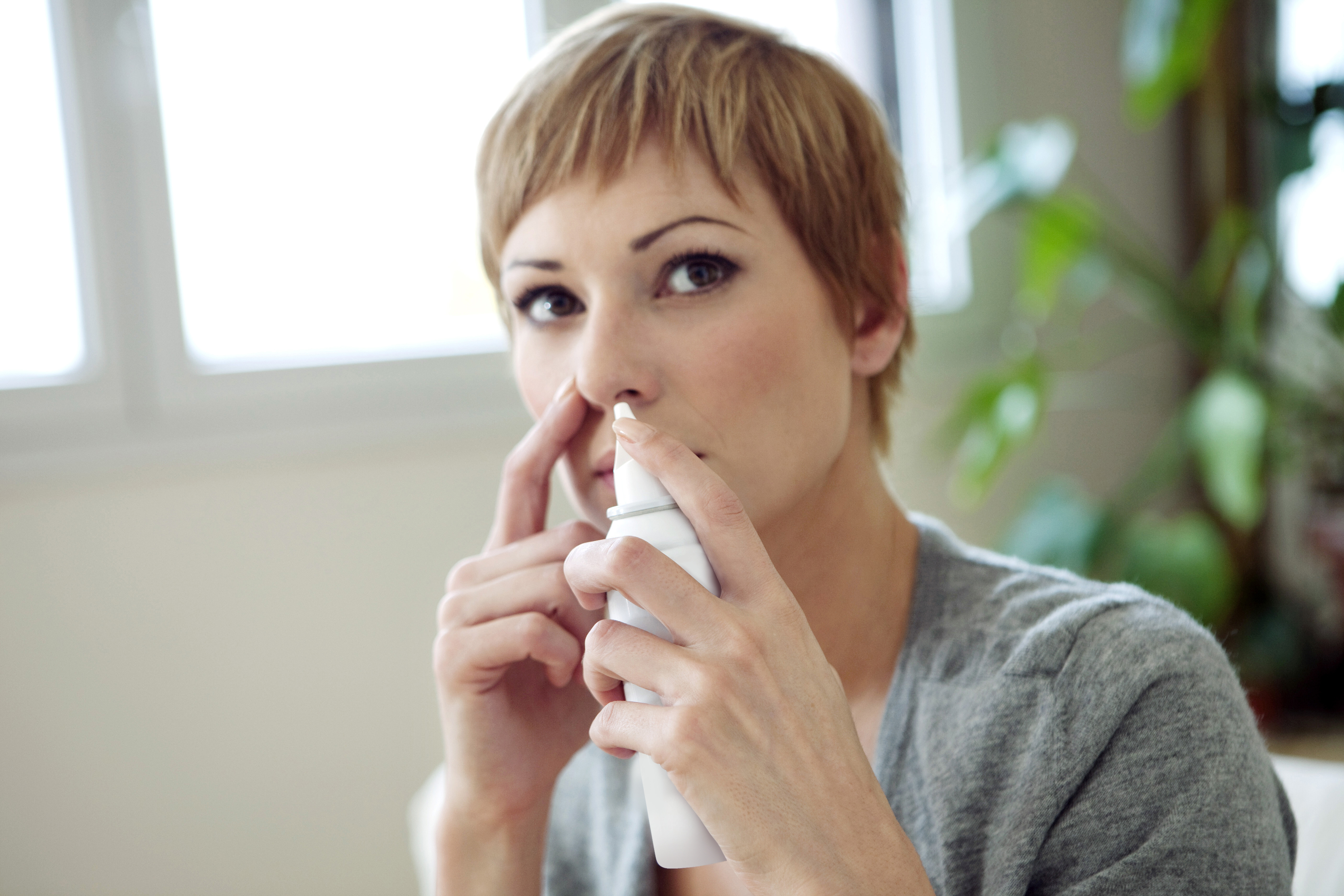 A woman with short blonde hair, wearing a gray sweater, uses a nasal spray with Aptar Pharma’s Bag-On-Valve technology, pressing the spray to her nose in a well-lit room with plants in the background.