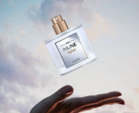 A hand is held out with a clear perfume bottle labeled INUNE by Aptar, featuring HDS INUNE a high definition spray fragrance pump, floating above it against a soft, cloudy sky background.