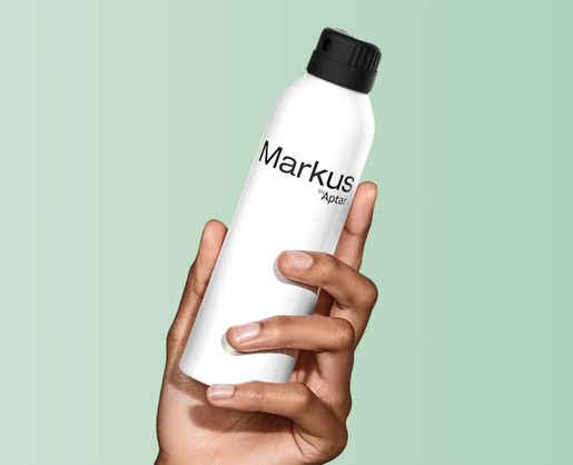 A hand holds a white spray can with a black premium twist to lock aerosol actuator against a light green background. The can is labeled Markus by Aptar.