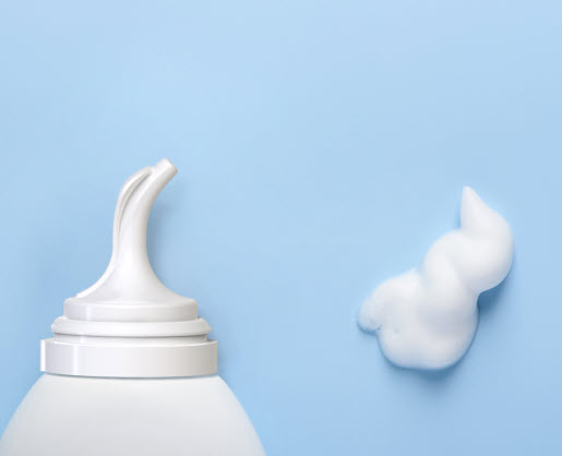 A close-up of a white dispenser named Athena bottle featuring an inverted aerosol actuator on a light blue background, with a swirl of white foamy product next to it.