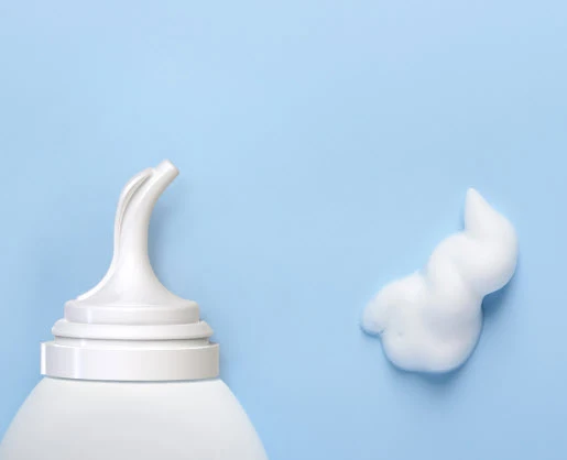 A close-up of a white dispenser named Athena bottle featuring an inverted aerosol actuator on a light blue background, with a swirl of white foamy product next to it.
