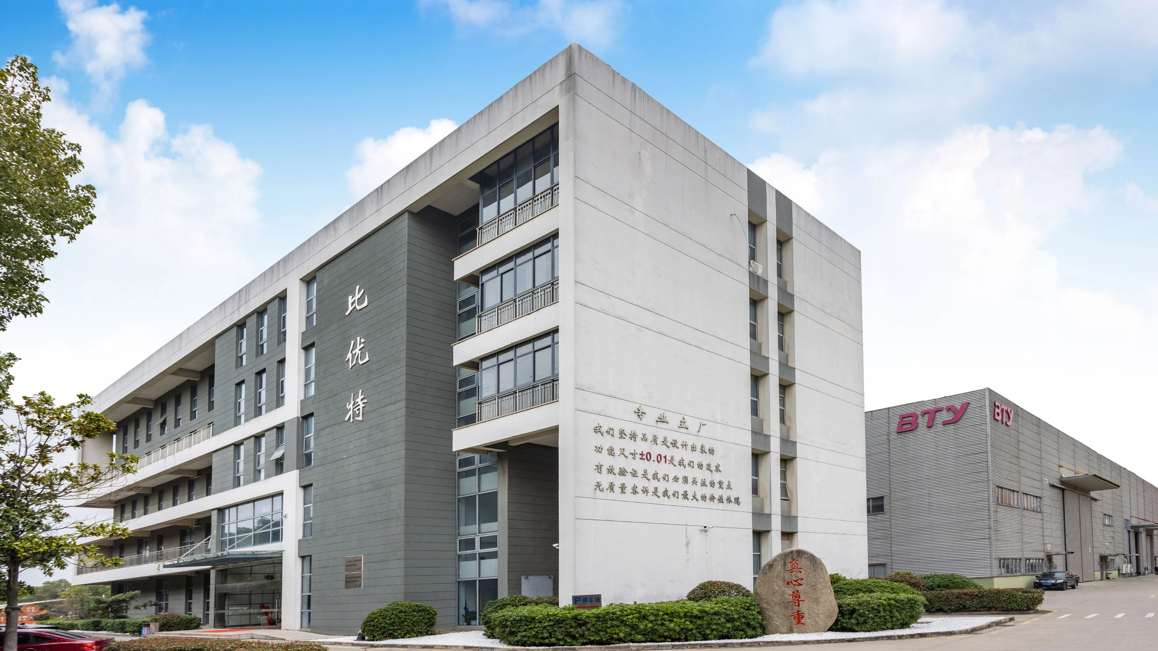 A picture of BTY's headquarters in Suzhou. A modern gray and white multi-story building with large windows and Chinese characters on its facade, next to a stone plaque and a separate industrial building labeled BTY under a blue sky with clouds.