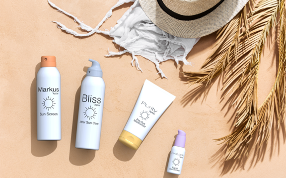 Set on a sandy surface with summer elements like a straw hat and palm leaves, four dispensing systems are showcased in a flat lay style including Markus, Bliss, Purity, and Evolock. The bottles have a 'sun' icon on them and the products are labeled with different types of suncare application such as sunscreen and post-sun after care.