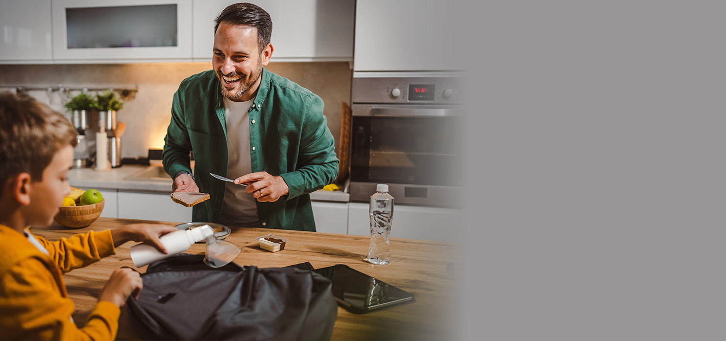 A young child in a brown shirt puts a white PureHale fine mist dispenser into a bag in family kitchen with his father.