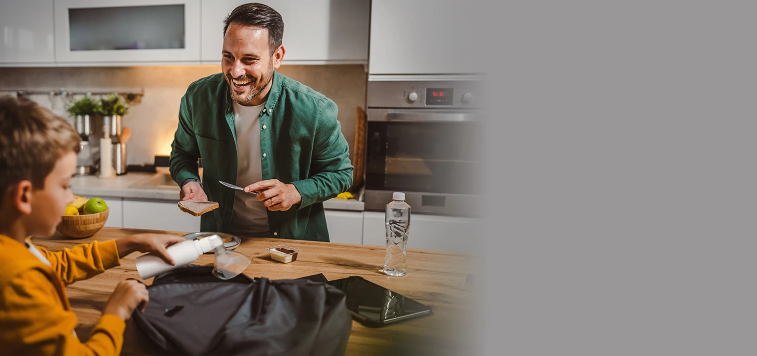 A young child in a brown shirt puts a white PureHale fine mist dispenser into a bag in family kitchen with his father.