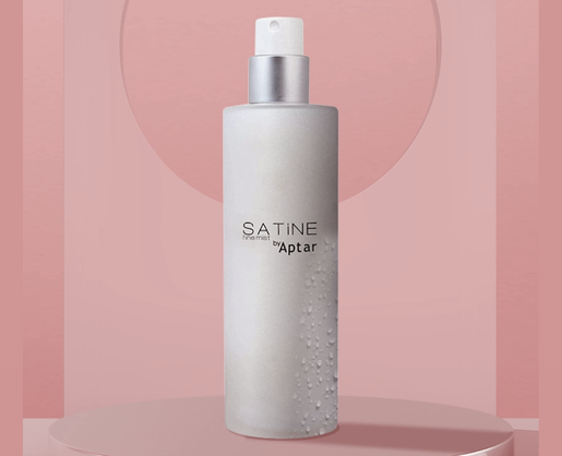 A frosted cylindrical spray bottle with a generous dosage cosmetic pump, named SATINE by Aptar, stands upright against a pink background.
