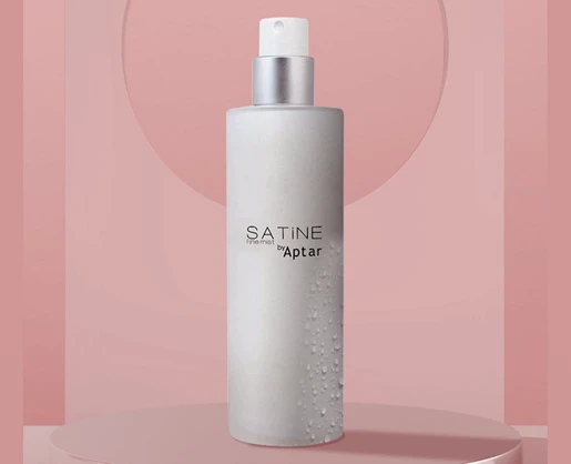 A frosted cylindrical spray bottle with a generous dosage cosmetic pump, named SATINE by Aptar, stands upright against a pink background.