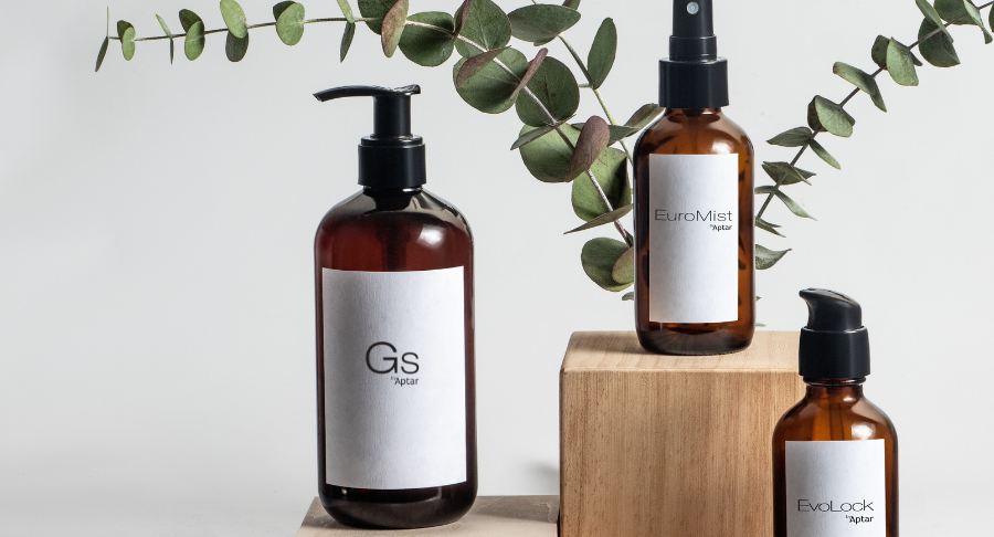 Three amber bottles labeled Evolock, Euromist, and GS, with GS featuring an Iconic High-Performing Dispensing Pump, displayed on and beside wooden blocks with a sprig of eucalyptus in the background against a neutral backdrop.