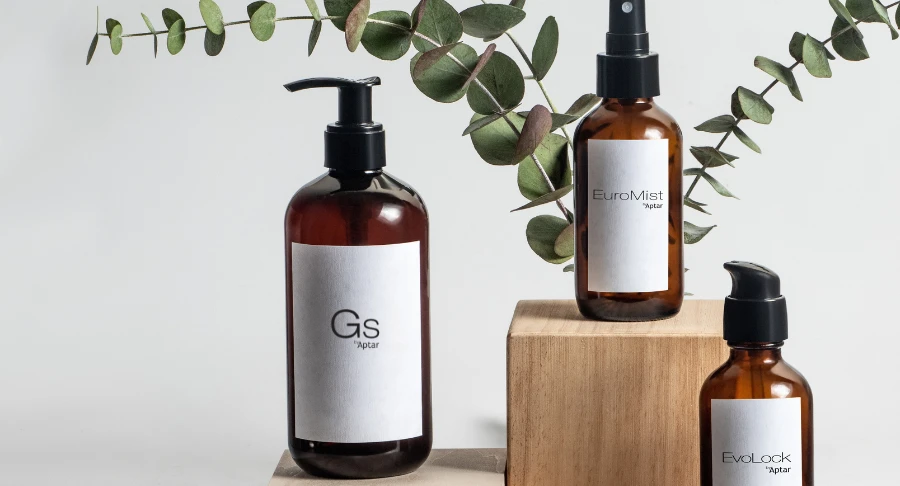 Three amber bottles labeled Evolock, Euromist, and GS, with GS featuring an Iconic High-Performing Dispensing Pump, displayed on and beside wooden blocks with a sprig of eucalyptus in the background against a neutral backdrop.