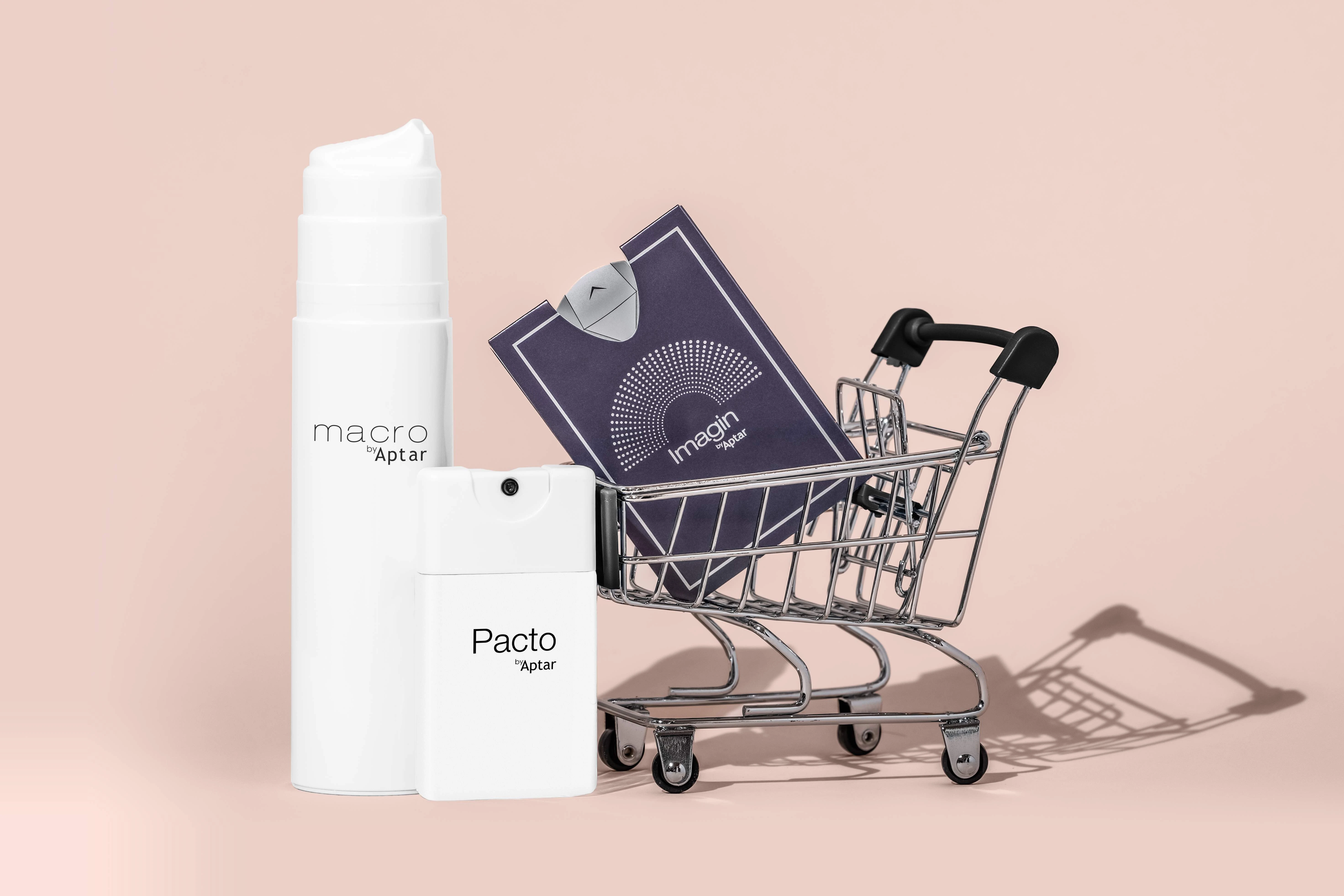 A small shopping cart holds a white Pacto by Aptar spray bottle, a purple Imagin which is an ultra-flat fragrance sampling, and stands next to a larger Macro airless packaging  all set against a light pink background.
