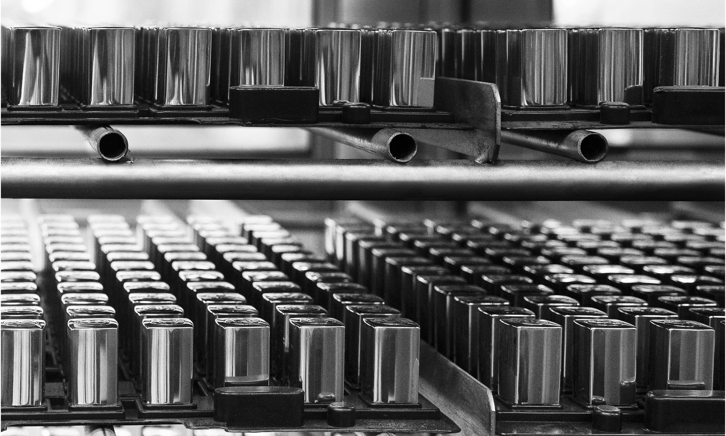 Rows of rectangular metal containers, possibly for a Non-Guided Lipstick Mechanism named Iconic, are neatly arranged on shelves in a factory or industrial setting. Their shiny surfaces create striking patterns in this black and white image.