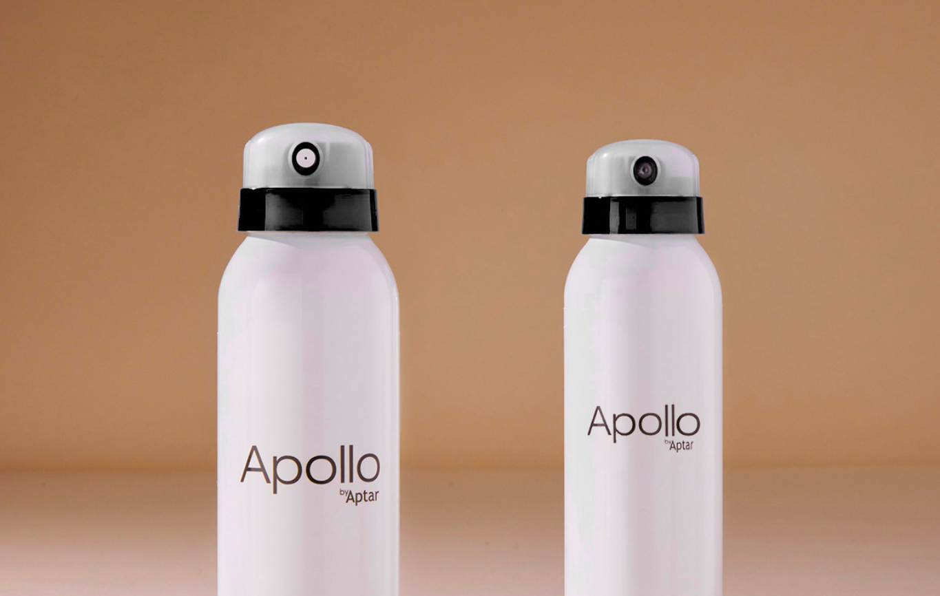 Two white spray bottles labeled Apollo by Aptar stand upright side by side against a plain beige background. Each bottle features a dual-spray aerosol actuator with  lightly grey-tinted clear cap and visible black and white spray nozzles. 
