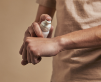 A person wearing a beige shirt applies a dollop of cream from a white luxury beauty dispensing named Bliss pump bottle onto the back of their hand, against a neutral background.