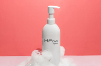 A white pump bottle labeled HiFlow by Aptar, featuring an e-commerce capable high-dose dispensing pump, stands amid white soap bubbles against a pink background.
