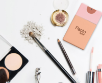 Assorted makeup items including brushes, eyelash curler, powder compact, broken eyeshadow palette, eyeliner pencil, lip gloss, and a peach-pink PACTO Fine Mist pocket size spray packaging by Aptar, all arranged on a white surface.