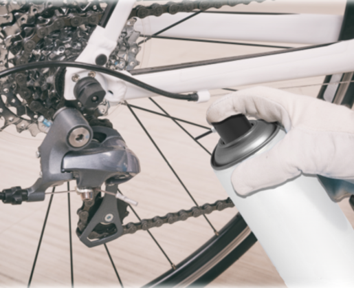 A gloved hand holds a spray can with Akur an aerosol actuator with built in tube near the derailleur and gear cassette of a bicycle, suggesting the bike is being cleaned or lubricated.