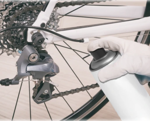 A gloved hand holds a spray can with Akur an aerosol actuator with built in tube near the derailleur and gear cassette of a bicycle, suggesting the bike is being cleaned or lubricated.
