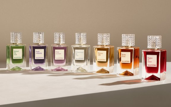 Seven classic Valentino perfume bottles, each with a different colored liquid and unique label, are arranged in a row on a light-colored surface, casting shadows in soft, warm lighting, a true display of prestige fragrance.