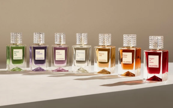 Seven classic Valentino perfume bottles, each with a different colored liquid and unique label, are arranged in a row on a light-colored surface, casting shadows in soft, warm lighting, a true display of prestige fragrance.