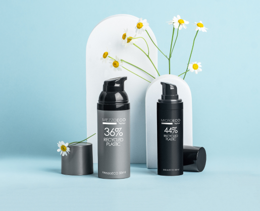 Two of Aptar Beauty's best-selling airless packaging labeled Mezzo, labeled 36% and 44% recycled plastic, stand on a light blue surface with white arch props and scattered daisies, set against a matching blue background. One bottle is full black with the cap placed next to it and the other is silver with a black actuator, and a silver cap placed next to it.