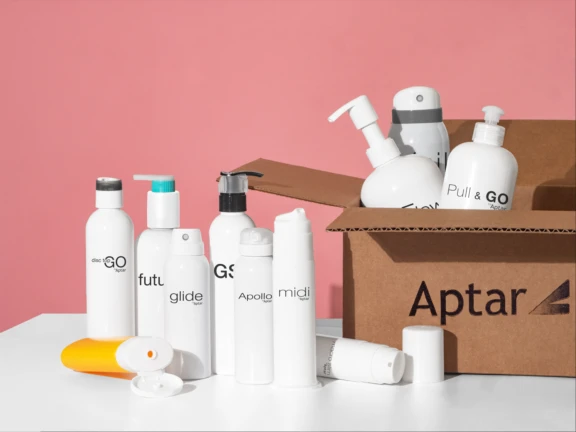 A group of white cosmetic bottles with various pumps and caps are arranged on a white surface next to a brown box labeled Aptar, showcasing stylish omnichannel packaging against a pink background. Some bottles are inside the box, others on the table.