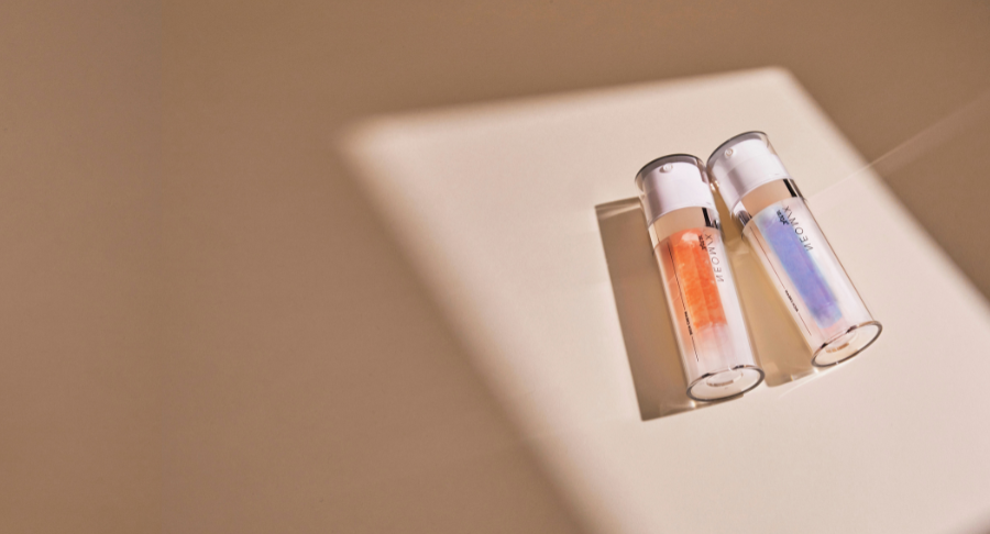 Two dual-chamber airless named Neomix skincare serum bottles with orange and blue liquid lie on a beige surface in soft, angled sunlight, casting long shadows.