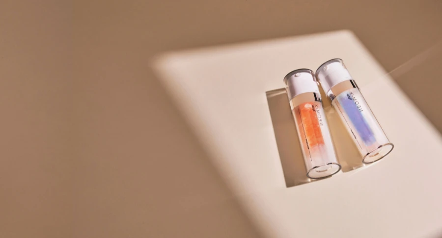 Two dual-chamber airless named Neomix skincare serum bottles with orange and blue liquid lie on a beige surface in soft, angled sunlight, casting long shadows.