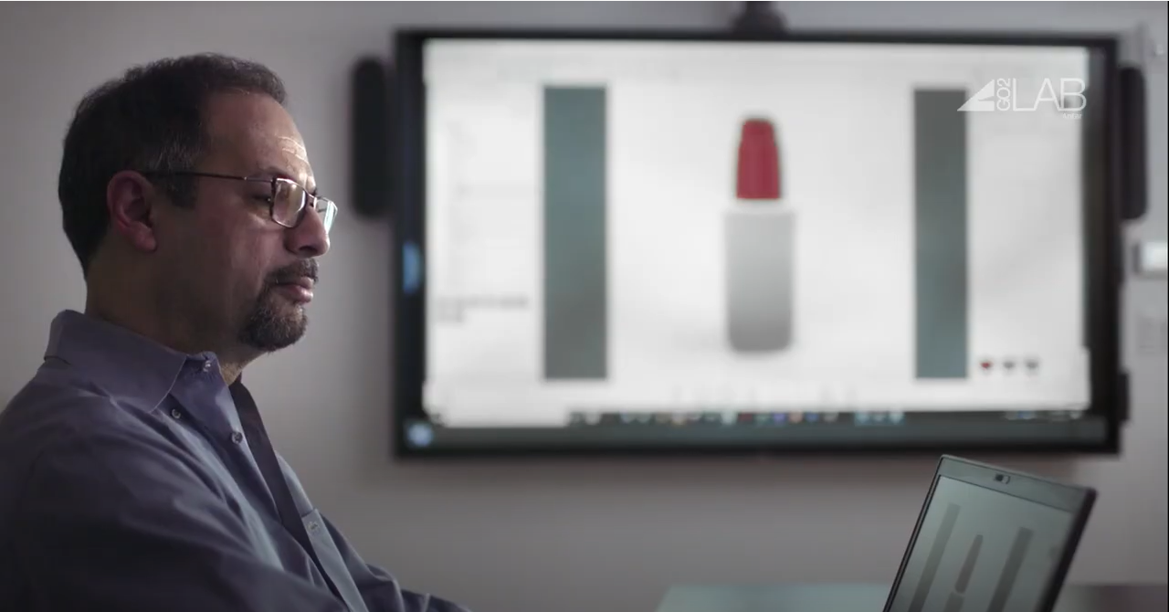 A man with glasses works on a laptop in an office. Behind him, a large screen displays a digital rendering of a lipstick and a vertical bar chart, highlighting the Aptar Go2Lab's professional and focused setting.
