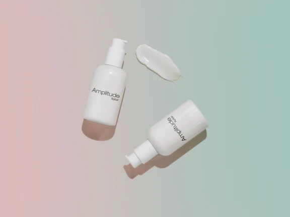 Two white Amplitude high dose cosmetic pumps on bottles sit on a soft gradient pastel background, with a smear of white cream near the top bottle.