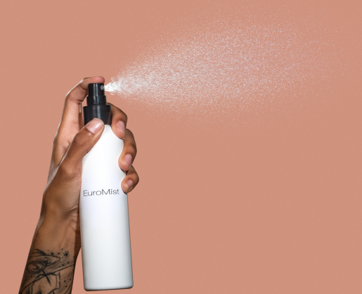 A hand with a tattoo uses a fine mist spray pump from a white bottle labeled EuroMist, releasing a delicate mist against a solid peach background.