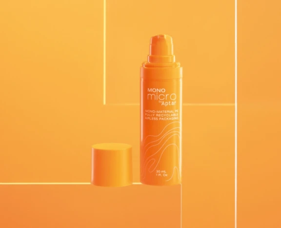 An orange airless pump bottle with its cap off, labeled “MONO micro by Aptar,” stands on an orange background with geometric light reflections. The bottle showcases fully recyclable, mono-material airless packaging.