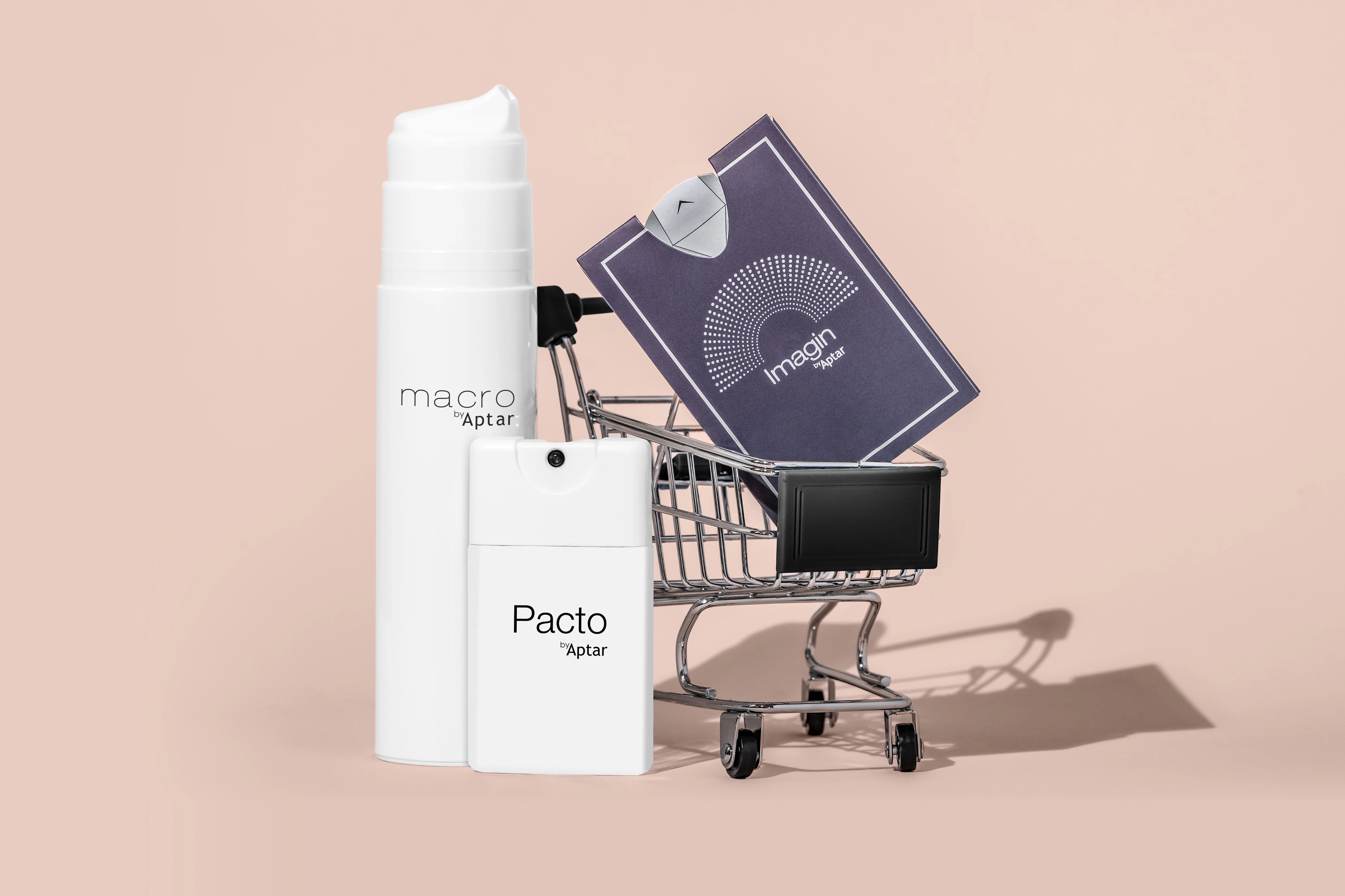 A small shopping cart holds a white bottle labeled macro Aptar, a pocket-size beauty packaging container labeled Pacto Aptar, and a purple booklet titled Imagin against a light beige background.